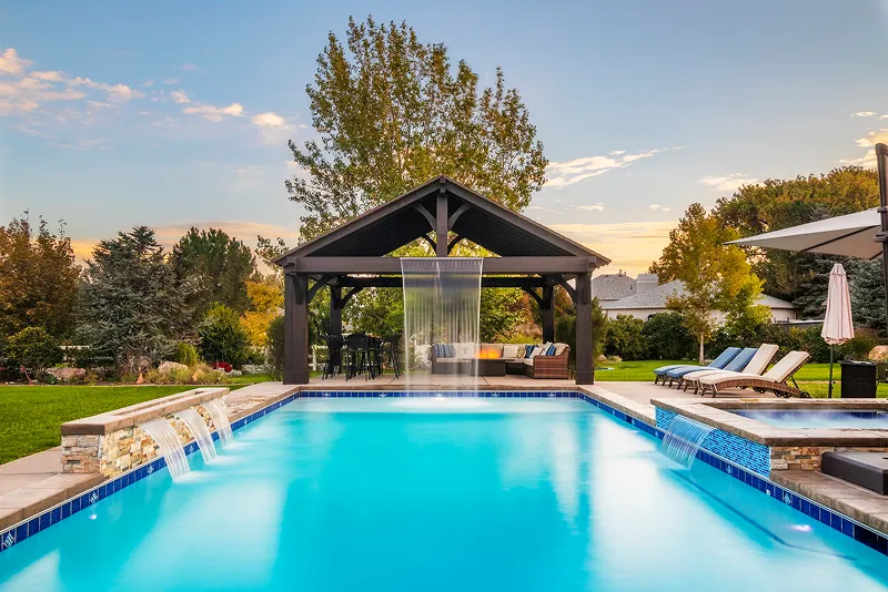 A dark wood pavilion with a sheer waterfall stands beyond an elevated hot tub and swimming pool on a patio at sunset.