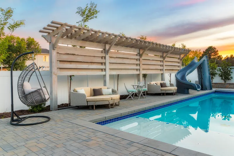An outdoor kitchen with a long bar and a separate sunken seating area with a fire pit are covered by a light wood pergola with string lights in a lush backyard.
