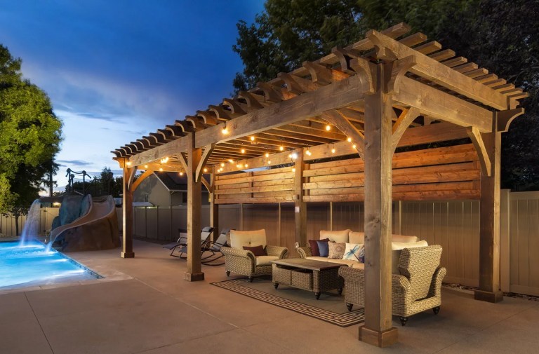 A beautiful backyard scene at dusk features a large rustic wooden pergola with string lights and horizontal wood privacy screens