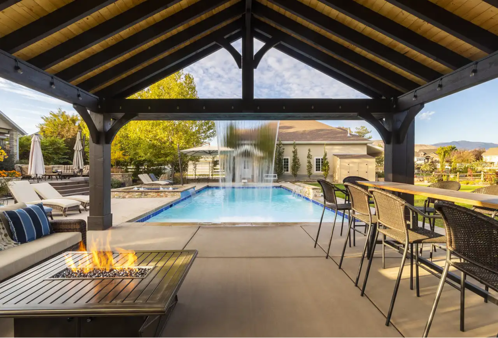 Looking out from under a dark-stained wooden pavilion with a light wood ceiling, a fire pit, bar, and seating face a swimming pool with a dramatic sheer descent waterfall.