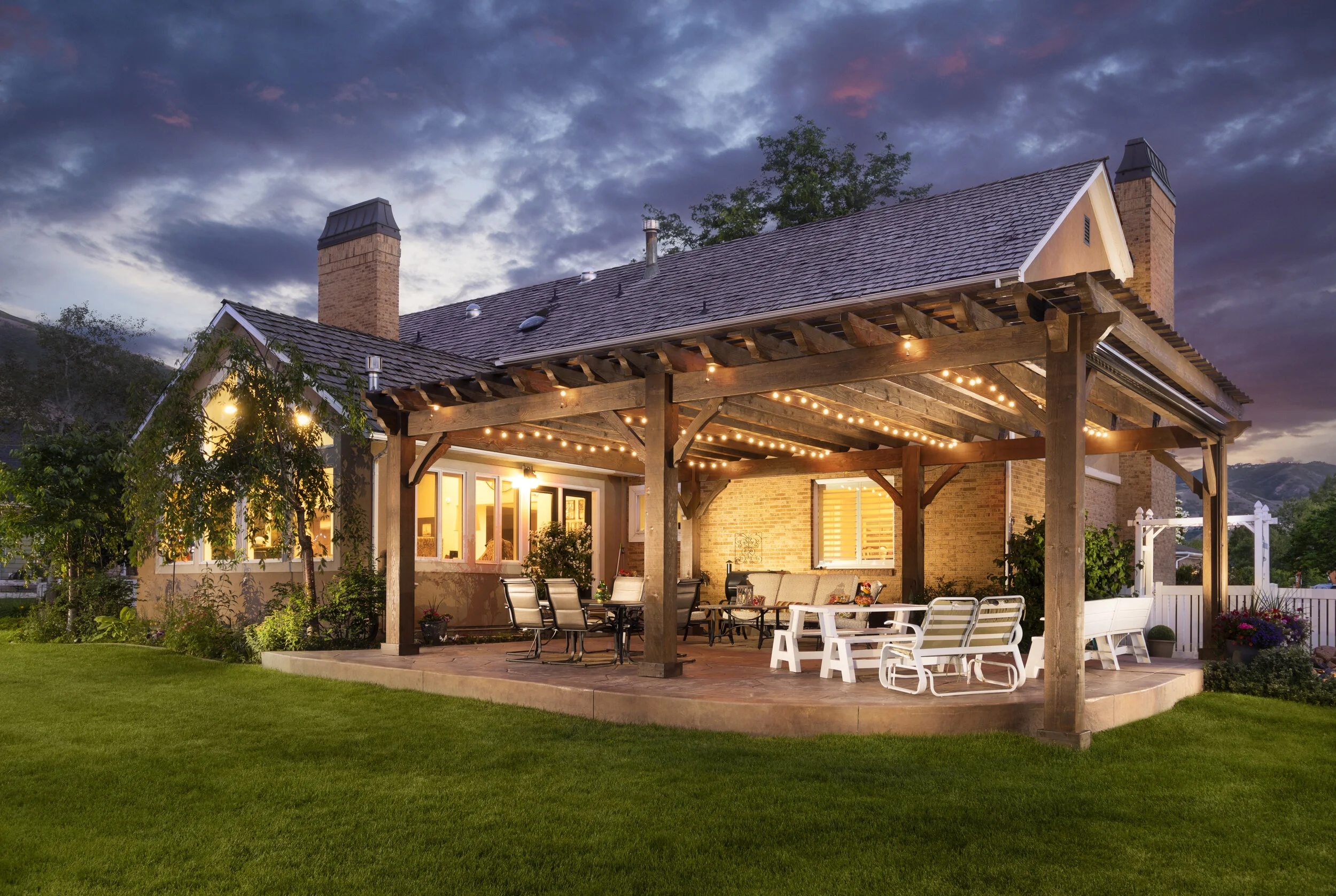 A rustic wood pergola with string lights covers an outdoor patio dining and seating area attached to a brick house at twilight.