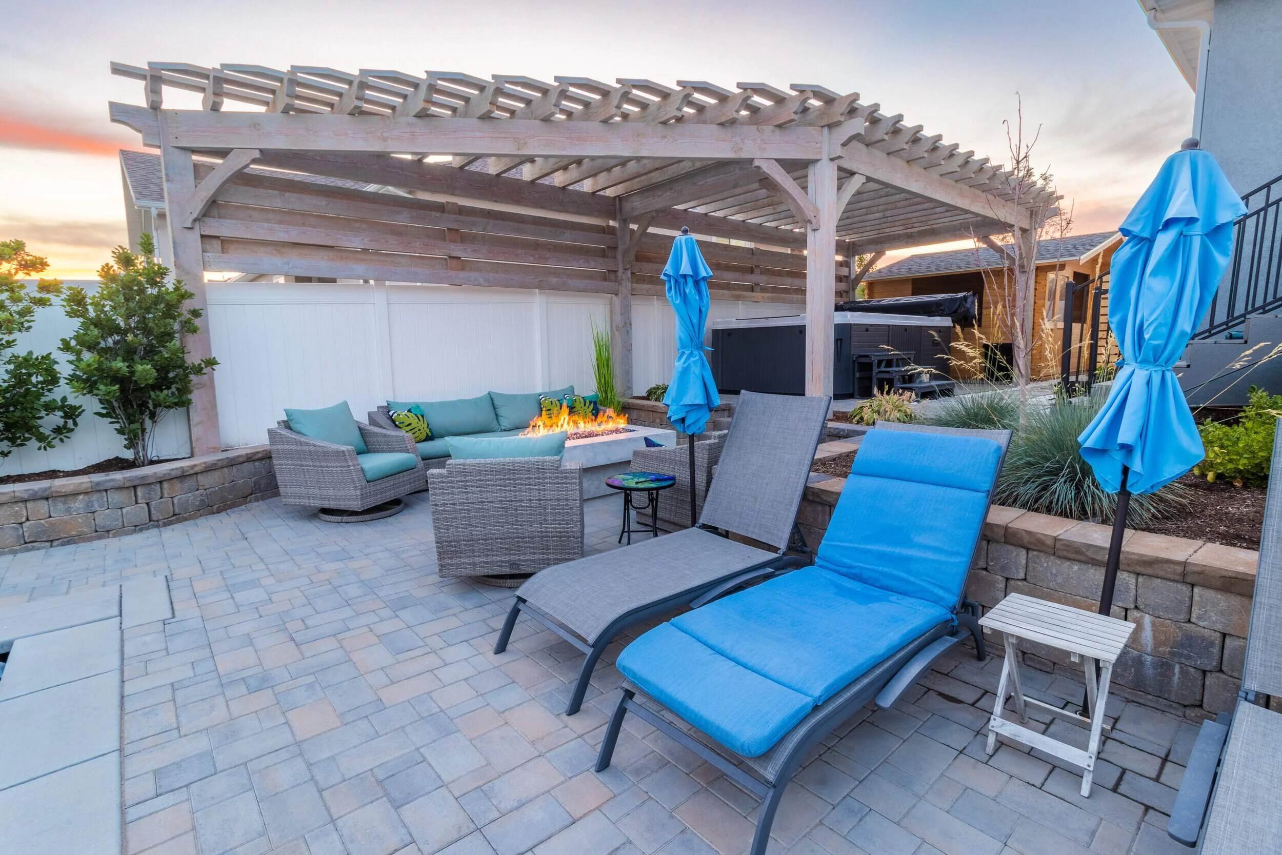 A large light-colored wooden pergola shelters a modern outdoor seating area with a fire pit and blue-cushioned furniture