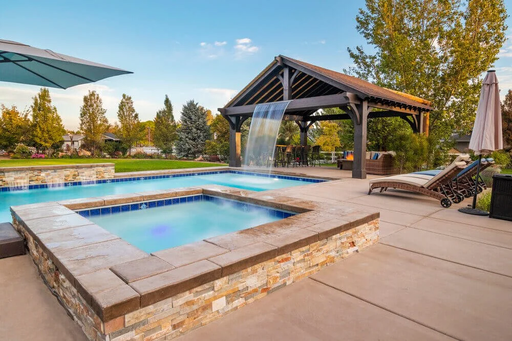 A dark wood pergola with a dramatic sheet waterfall stands behind an elevated hot tub and swimming pool on a concrete patio at sunset.