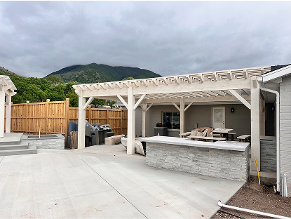 A white-washed wood pergola covers an outdoor kitchen and bar with a gray stacked-stone façade on a new concrete patio