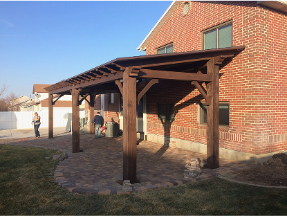 A newly constructed, dark-stained wooden pergola with a slanted roof stands over a circular paver patio attached to a red brick house