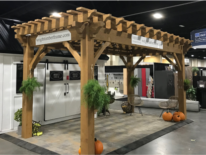 A medium-brown wooden pergola with company logos is displayed on a paver floor as part of a trade show booth