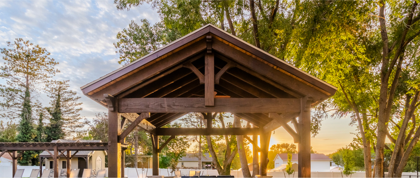 A dark-stained wooden pavilion with a peaked roof stands in a backyard