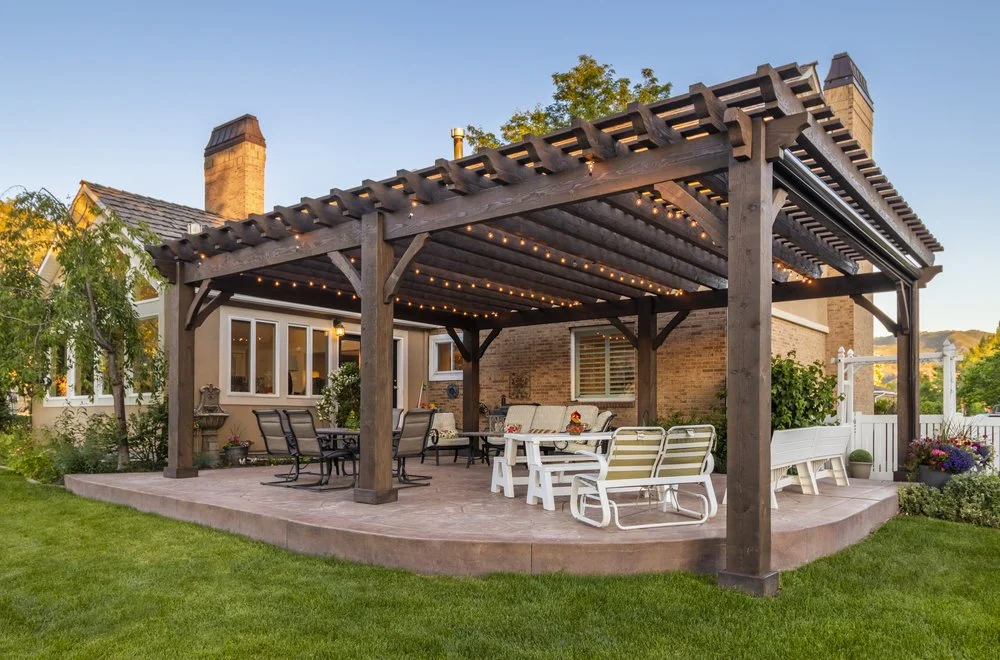 A large, dark-stained wooden pergola with string lights covers an outdoor patio furnished