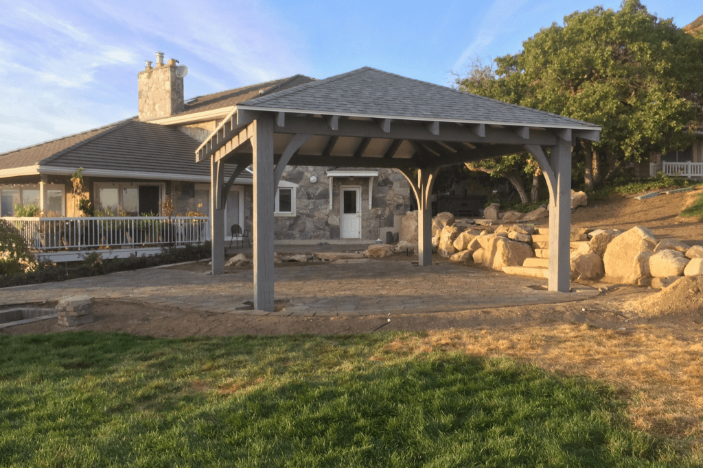 A large, light gray wooden gazebo with a hip roof sits on a paver patio being constructed in front of a stone-clad house and a rock retaining wall.