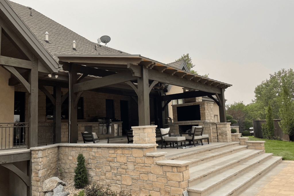 An exterior view of a large, luxurious covered patio featuring a stone wall, outdoor furniture, a fireplace, and a mounted television.
