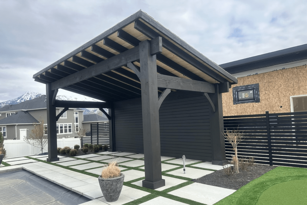 A large dark-stained wooden pavilion sits on a modern, gridded paver patio next to a covered swimming pool and a horizontal slat fence.