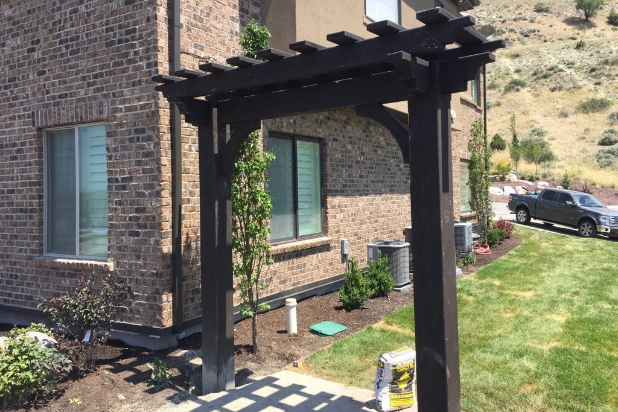 A dark-stained wooden pergola acts as an entrance or archway between a brick house and a grassy yard