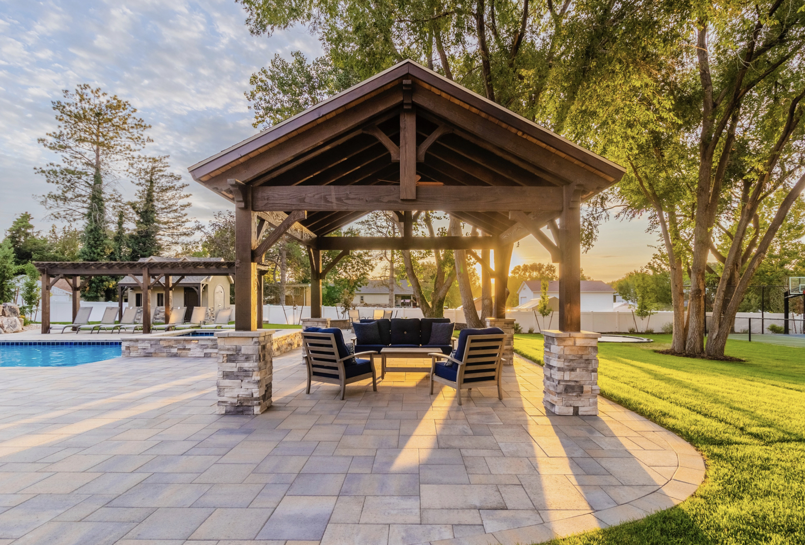 Timber Frame Pavilion by Poolside at Sunset Dark timber frame pavilion with stone base columns and outdoor seating, overlooking a pool and landscaped backyard at sunset