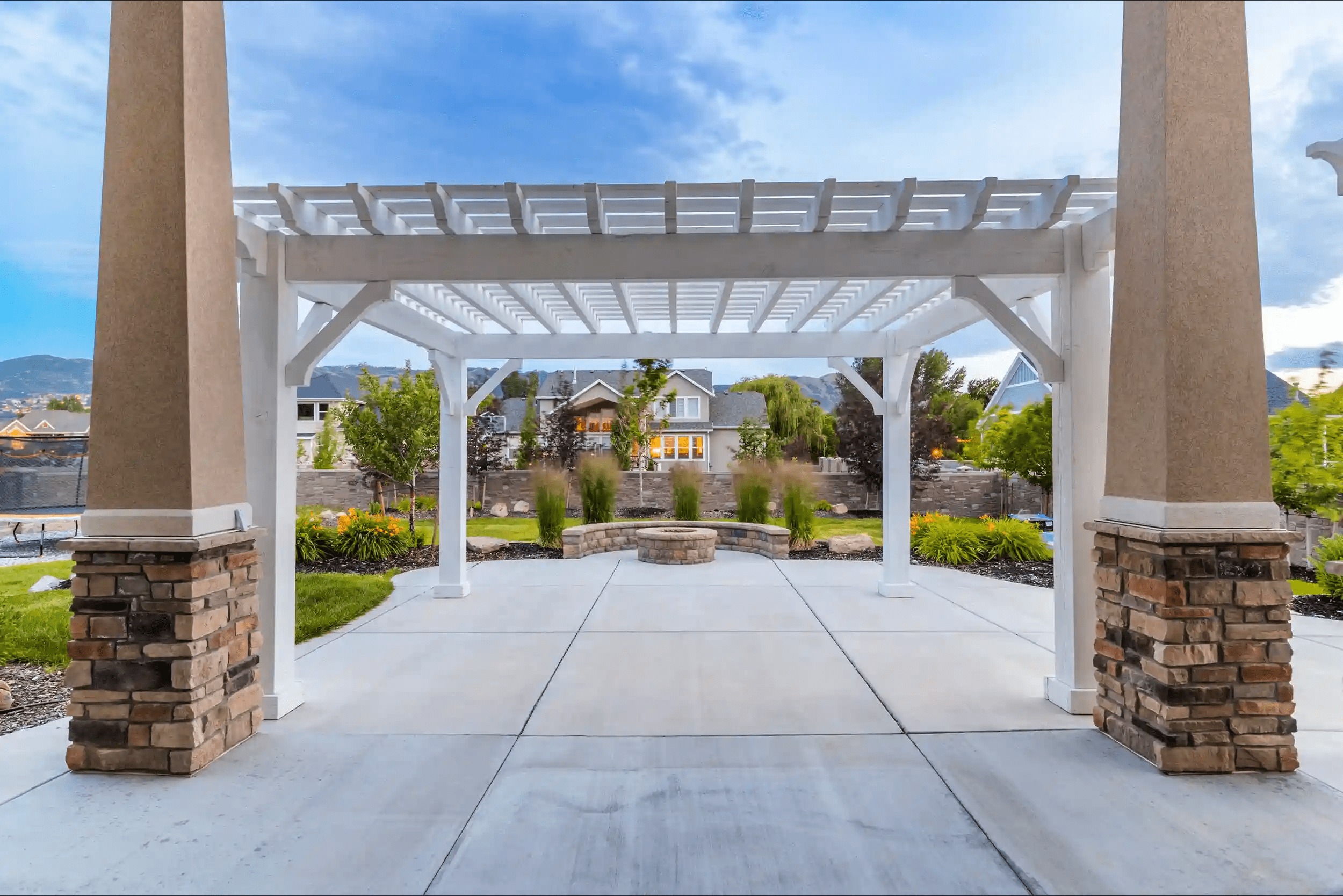 Elegant White Timber Frame Pergola with Patio Open-lattice Timber frame pergola in white over a patio with stone columns, framing a fire pit and landscaped yard