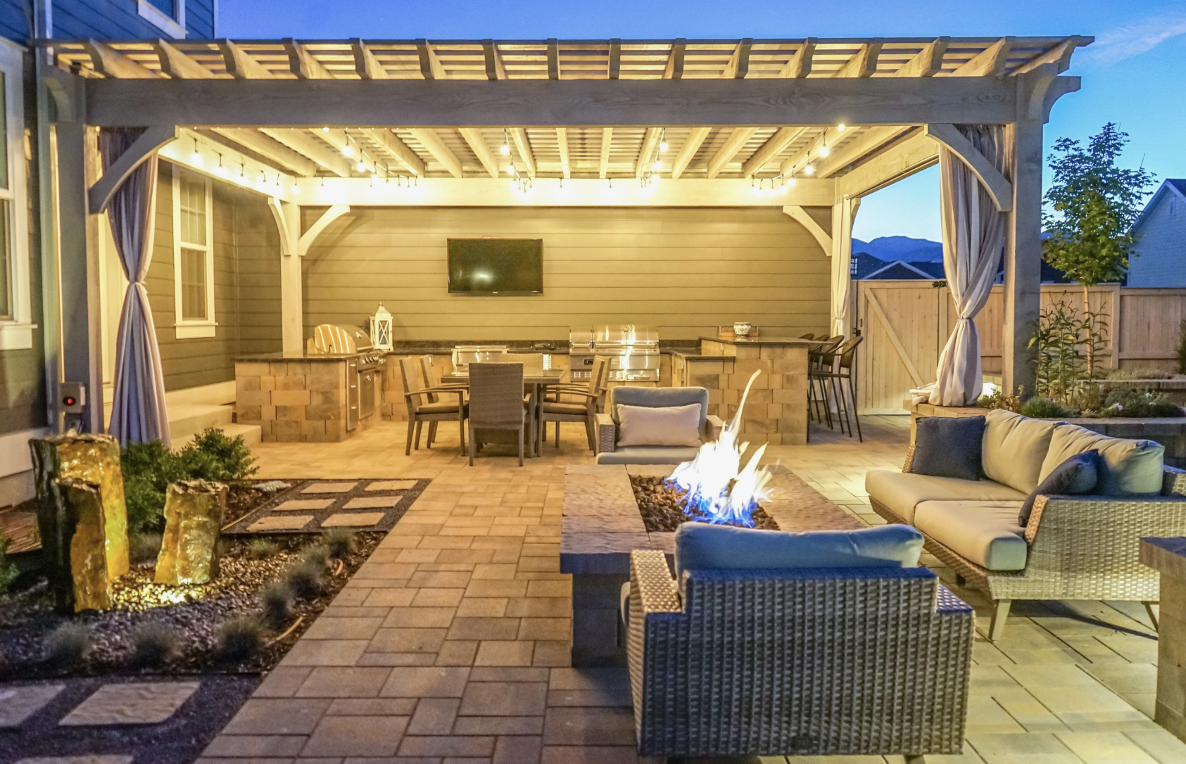 Evening patio scene with a custom pergola for outdoor living, string lights, outdoor kitchen, and seating by a fire pit