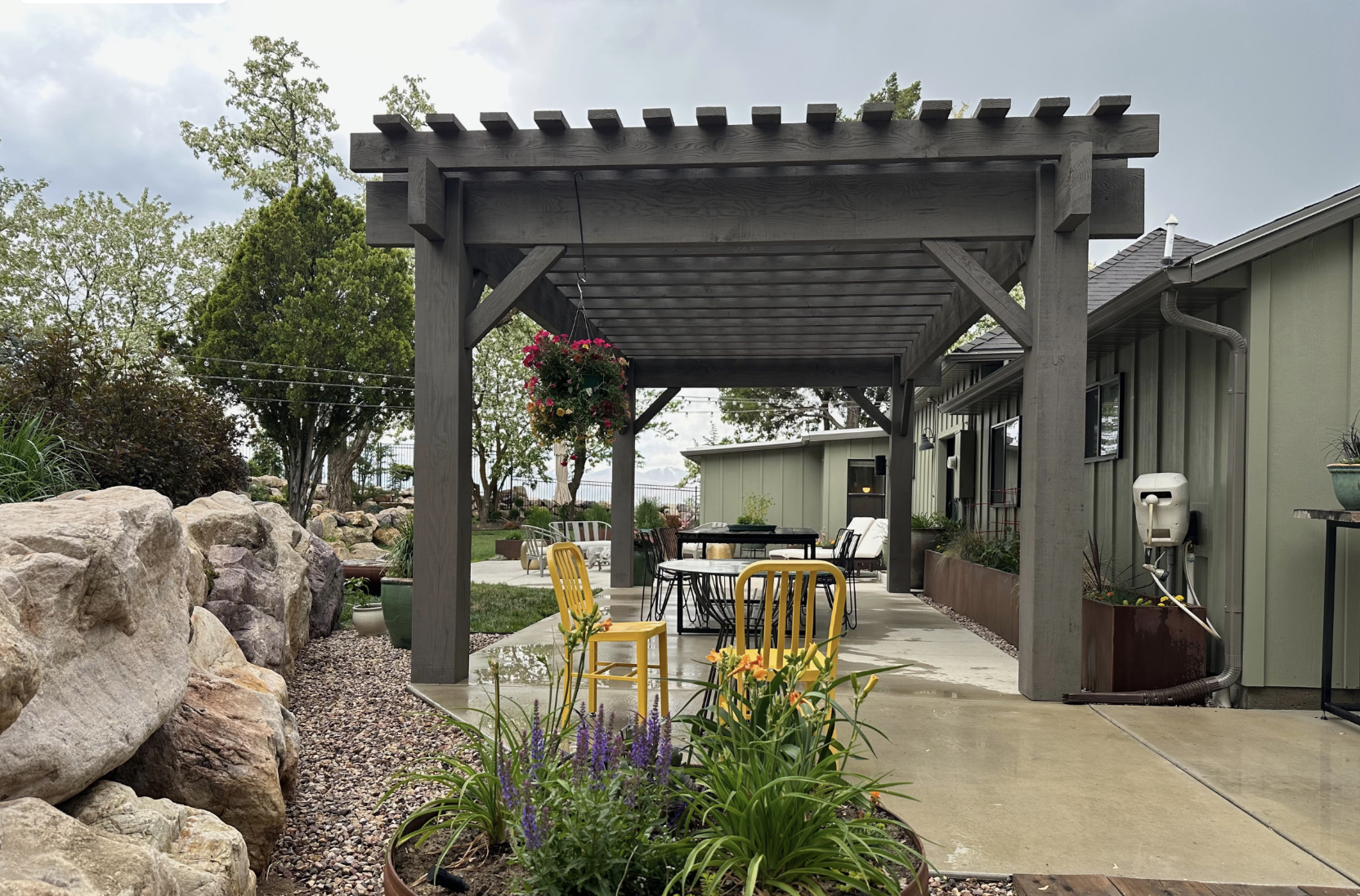 Modern Backyard Pergola with Cozy Patio Seating Gray timber backyard pergola over a concrete patio with dining area, hanging basket, and blooms along a rock border