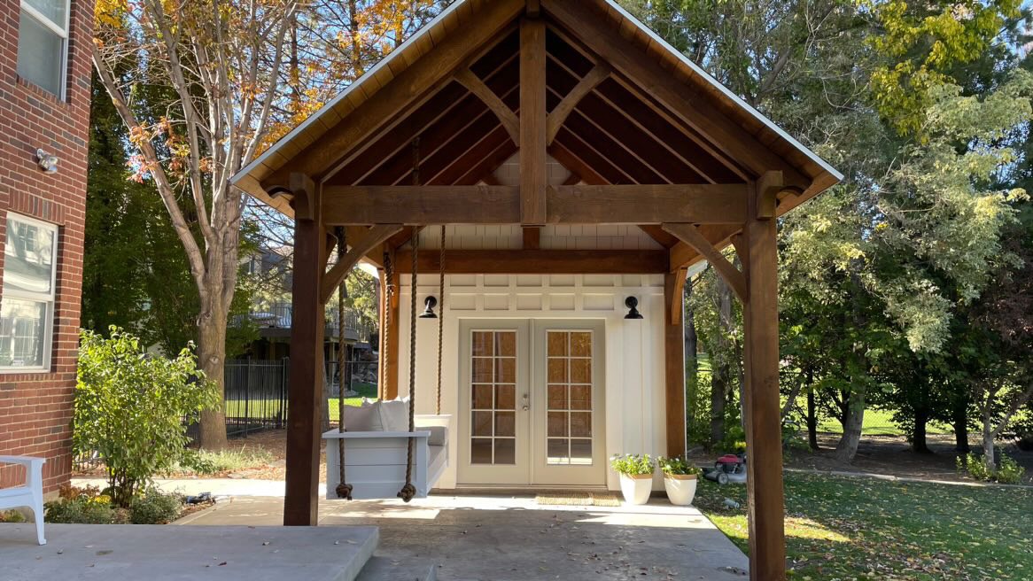 A cozy pavilion attached to house featuring exposed wooden beams, a hanging bench swing, and French doors opening to a garden view.