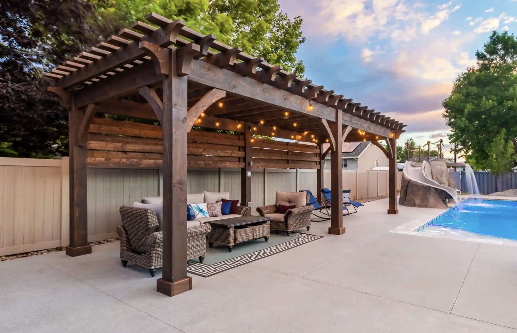 Covered backyard pergola with cozy seating by a pool, showcasing the best time to buy a pergola for outdoor living.