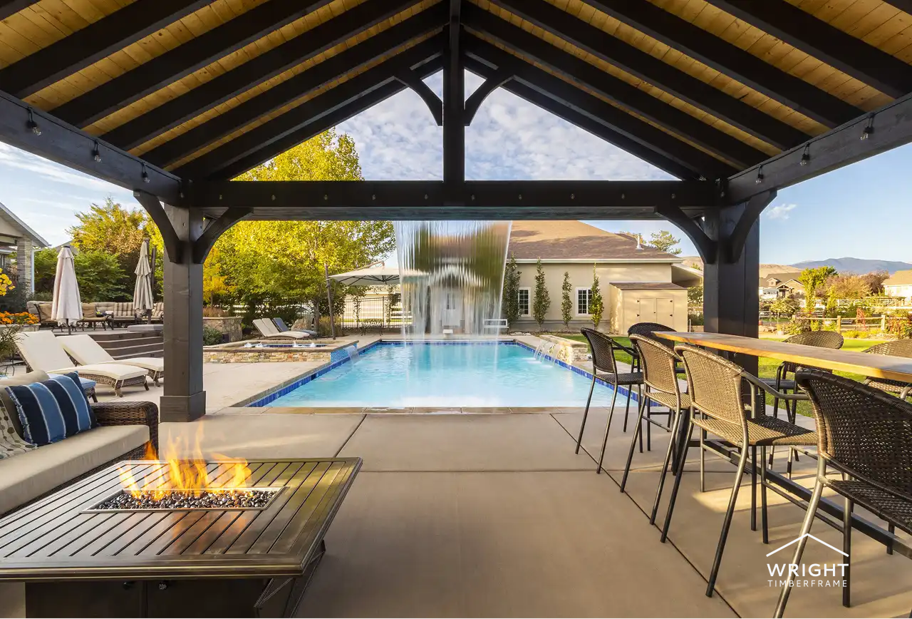 Modern timber frame pavilion with a fire table, overlooking a pool and waterfall, featuring comfortable seating and outdoor dining space, enhancing outdoor living aesthetics.